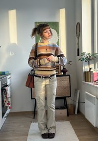 Pull rayé dans des tons bruns et bleus, pantalon cargo beige, chaussures marron et sac porté en bandoulière marron. Intérieur avec un sol en bois clair.