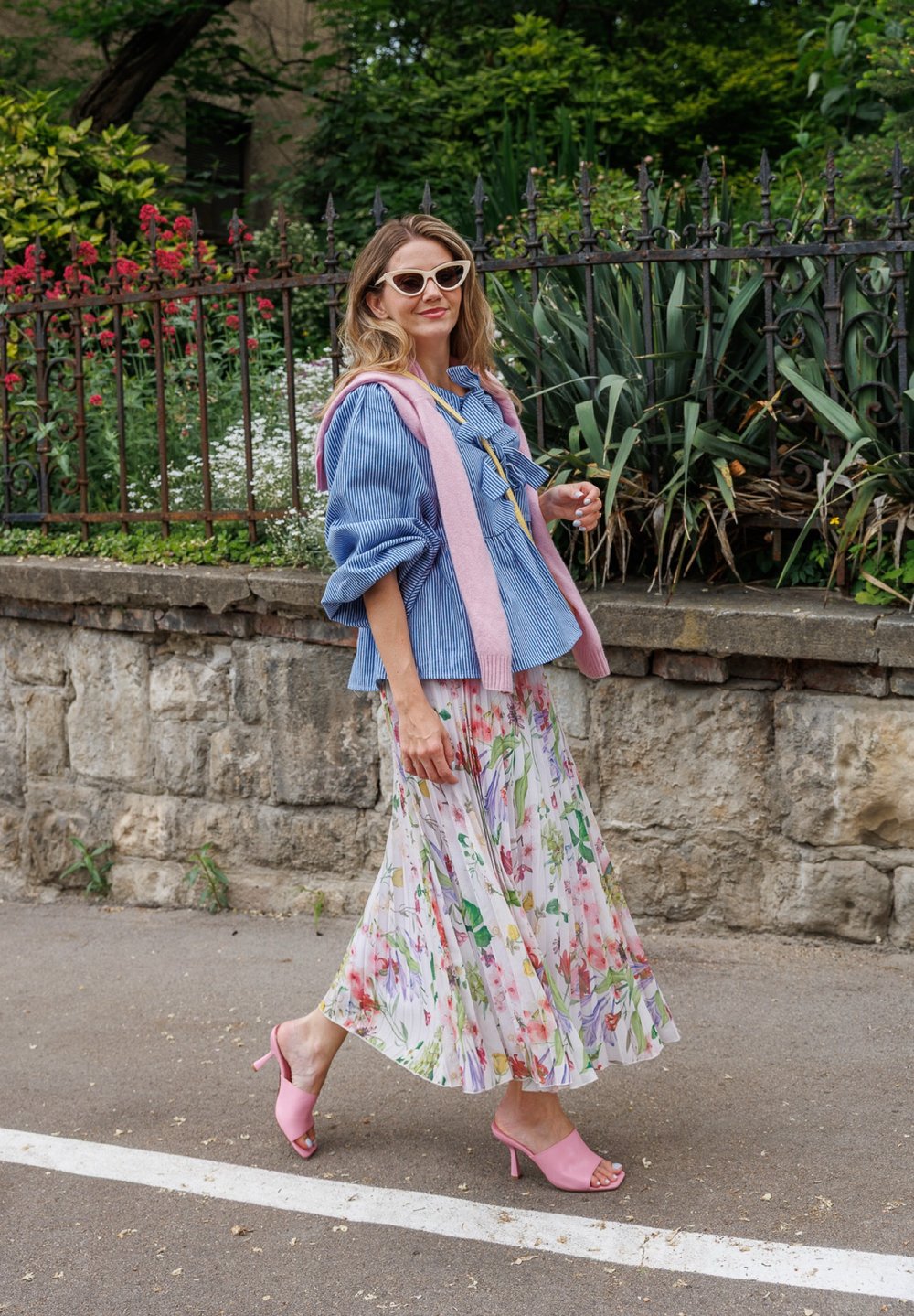 Blusa blu a righe con maniche a palloncino, gonna maxi fiorata in tonalità pastello, maglione rosa drappeggiato e sandali rosa con apertura sulla punta.