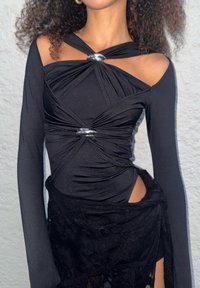 Svart langermet body med vridd design, sølvdetaljer og utskjæring ved midjen, laget av et glatt, elastisk materiale.