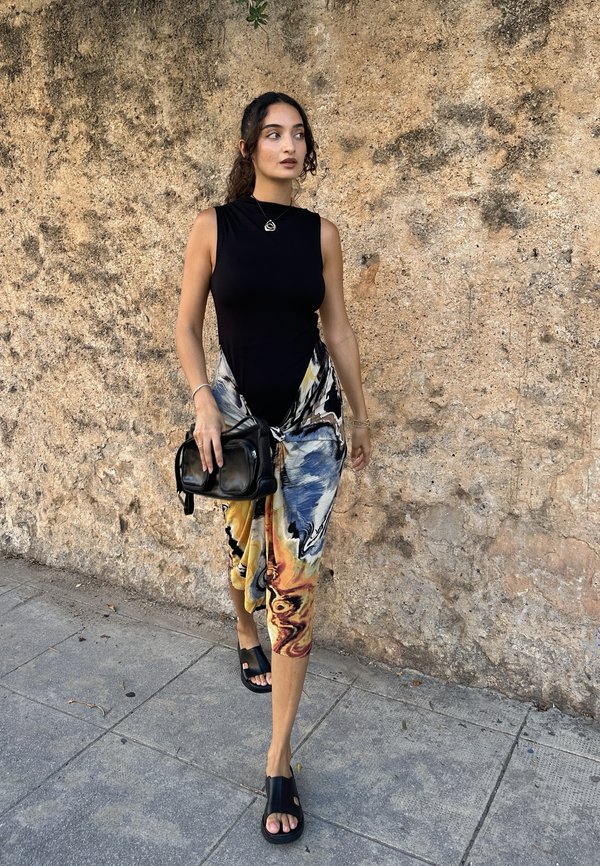 Femme marchant sur un trottoir portant un haut noir sans manches, une jupe colorée à motifs, des sandales noires et portant un sac à main noir.