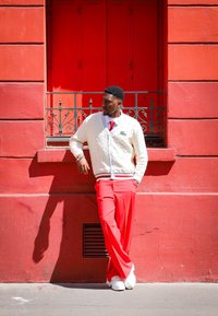 Λευκό πλεκτό πουλόβερ με μπλε ρέλι, κόκκινες παντελόνες και λευκά παπούτσια μπροστά σε έναν κόκκινο τοίχο, δίπλα σε ένα παράθυρο με διακοσμητικά σιδερένια στοιχεία.