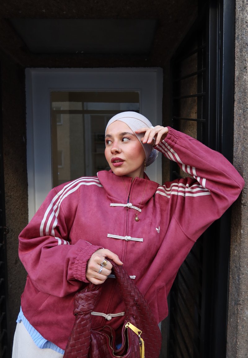 Jeune femme portant un foulard blanc et une veste rouge, tenant un sac à main tissé rouge, debout dans une porte regardant sur le côté.