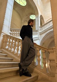 Mężczyzna w czarnym golfie i szarych szerokich spodniach stoi na schodach z ozdobnymi białymi balustradami i szczegółowymi cechami architektonicznymi.