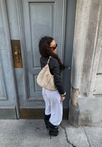 Bolsa de ombro em camurça bege com um design descontraído e forma curvada. Calças de fato cinzentas e botas pretas até ao joelho completam o visual.