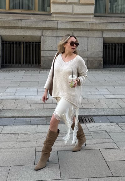 Mujer caminando por la acera de la ciudad con un suéter blanco, falda con encaje, botas marrones, gafas de sol y sosteniendo una bebida con pajilla.