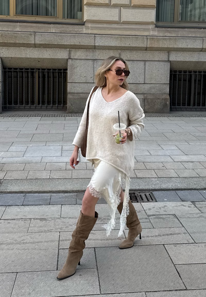 Frau läuft auf dem Bürgersteig in der Stadt, trägt einen weißen Pullover, einen Rock mit Spitzenbesatz, braune Stiefel, Sonnenbrille und hält ein Getränk mit einem Strohhalm.