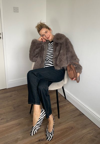 Mujer sentada en una silla, vestida con una chaqueta de piel gris, camisa a rayas blancas y negras, pantalones negros, zapatos con estampado de cebra y sosteniendo un bolso marrón.