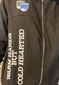 Μαύρο τζάκετ με φερμουάρ που διαθέτει δύο μπλέ και λευκές ρέλιες στο στήθος και λευκό κεντητό κείμενο στο μανίκι. Μαλακή υφή υφάσματος.