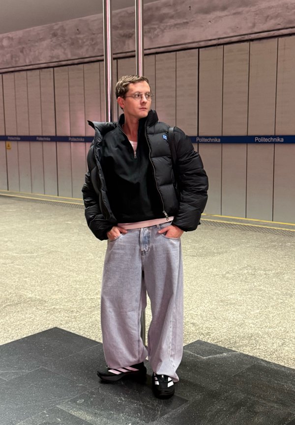 Chaqueta acolchada negra con cremallera sobre una camiseta negra; jeans de pierna ancha en gris claro; zapatos negros con detalles en rosa. Ambiente urbano interior.