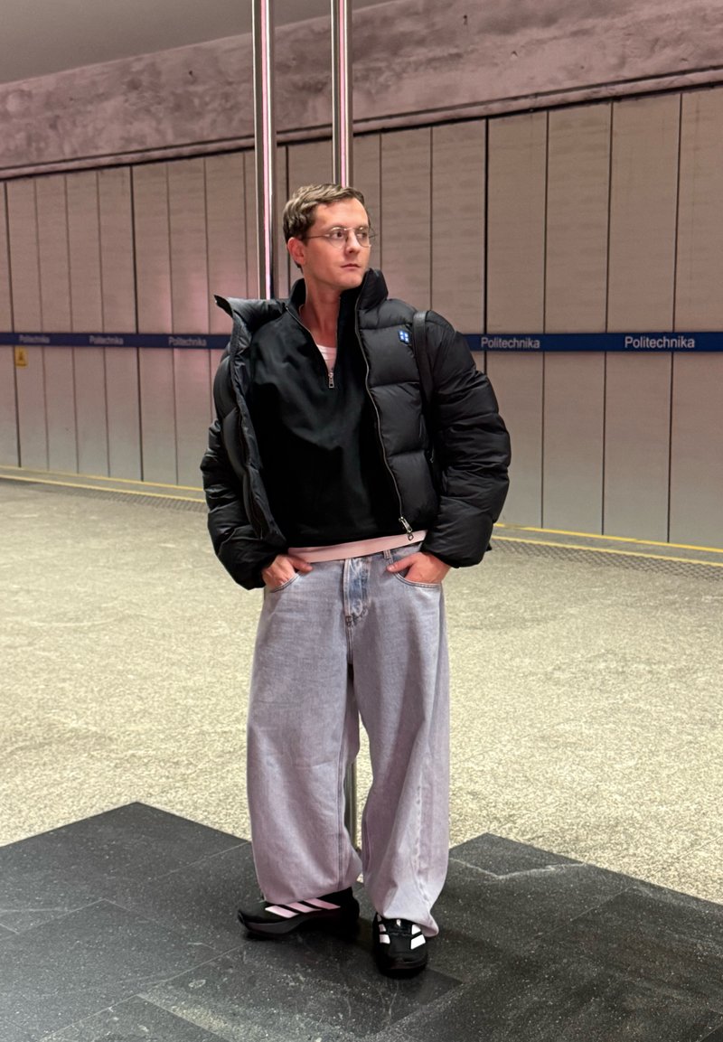 Doudoune noire zippée, portée sur un haut noir ; jean large gris clair ; chaussures noires avec des accents roses. Décor urbain intérieur.