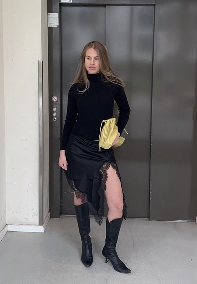 Joven mujer con jersey de cuello vuelto negro, falda negra asimétrica con encaje, botas negras hasta la rodilla, sosteniendo un bolso amarillo, de pie frente a un ascensor cerrado.