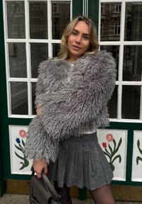 Sivá bunda z umelého kožúšku so strapatou textúrou, doplnená plisovanou sivou sukňou. Modelka drží tmavú kabelku na zeleno-bielom pozadí.