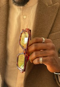 Hand wearing two silver chain-link rings holding amber-tinted rectangular glasses, dressed in a beige shirt and a brown wool coat.