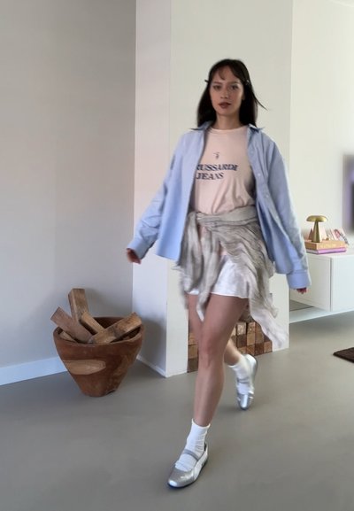 Una modelo camina con una camisa abotonada azul claro, una camiseta gráfica rosa, una falda blanca y zapatos plateados, con troncos de madera en una maceta de cerámica cerca.