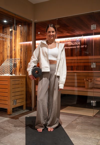 Mujer con ropa deportiva informal sosteniendo una pesa, de pie descalza sobre una esterilla frente a una sala de sauna con paredes de vidrio.