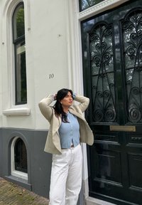 A light beige blazer is worn over a light blue knitted top, paired with white patterned trousers, standing against a dark green ornate door with window accents.
