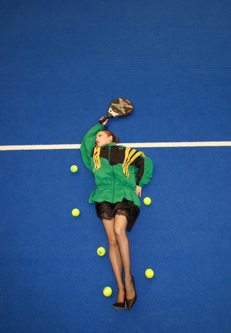 Donna con giacca verde e gonna nera sdraiata su un campo da tennis blu che tiene in mano una racchetta da padel, circondata da palline da tennis sparse.