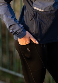 Mustad jooksutrikotid küljetaskuga, millel on hallis toonis brändi logo. Sobitatud sinise kerge jopega. Nähtav tekstuuriga kangas.