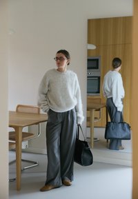 Svijetlosivi, puhasti pulover; labave sive hlače; smeđi papuče; drži crnu kožnu torbu; moderna kuhinja s drvenim akcentima.
