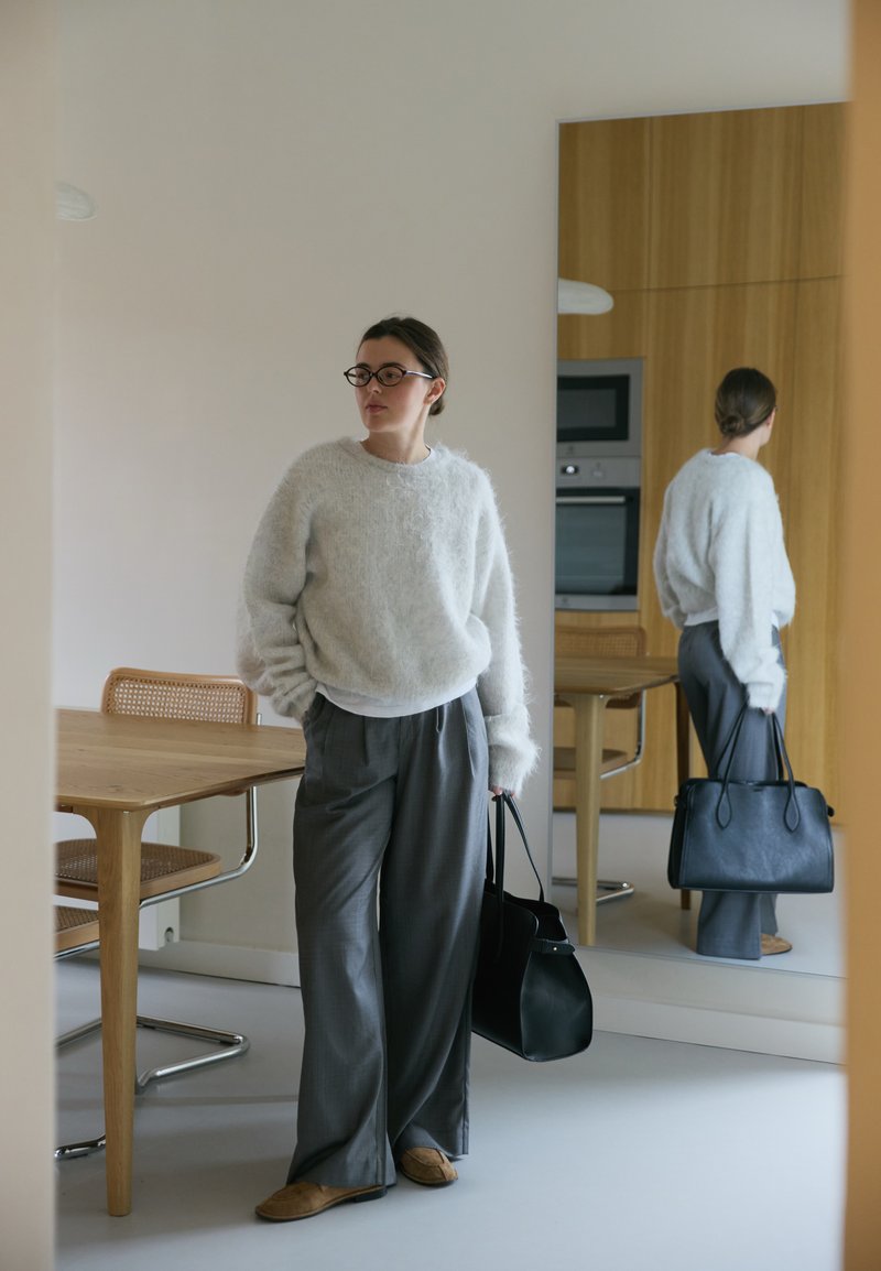 Helles graues, flauschiges Sweatshirt; lockere graue Hosen; braune Hausschuhe; hält eine schwarze Ledertasche; moderne Küche mit Holzakzenten.