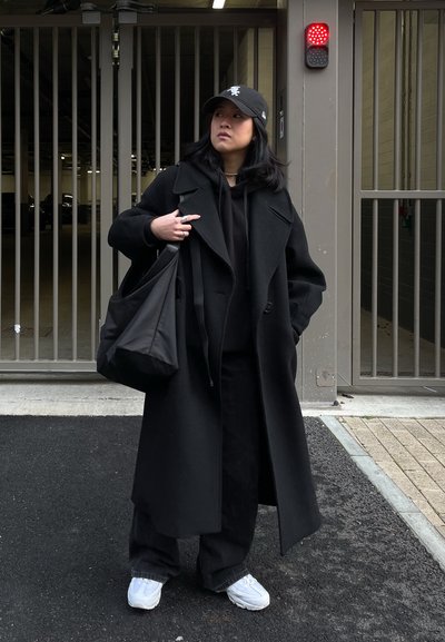 Abrigo de lana negra con solapas anchas, superpuesto a una sudadera negra con capucha. Combinado con pantalones negros holgados y zapatillas blancas. Llevando una bolsa tote negra.