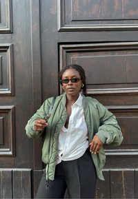 Giubbotto bomber verde con polsi a costine, camicia bianca e pantaloni neri. Gli accessori includono occhiali da sole sottili e rettangolari e collane a strati.