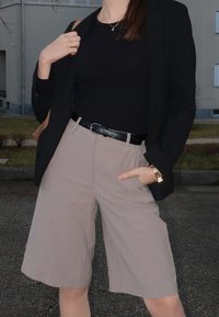 Personne portant un blazer noir et un haut, un short beige à jambes larges, une ceinture noire, une montre en or et un collier, debout à l'extérieur près d'un bâtiment.