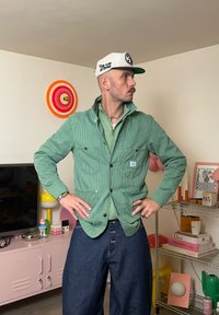 Grønn stripete jakke med to framlommer, lagd over en grønn genser med glidelås, kombinert med mørkeblå jeans. Hvit caps med logo.