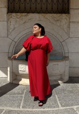 Vestido maxi rojo con mangas cortas abullonadas, tejido texturizado y un corte holgado. Lleva sandalias negras y está de pie junto a una fuente de piedra.