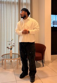 Homme portant une chemise blanche rayée, un pantalon noir et une casquette, debout à l'intérieur près d'une table ronde en verre avec un vase de fleurs et une chaise marron.