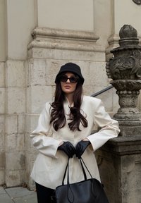 Blazer blanc à manches longues, chapeau seau en tricot noir, lunettes de soleil noires, gants noirs et sac à main noir, le tout sur fond de mur en pierre.