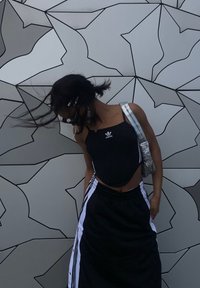 Débardeur noir court avec logo Adidas, pantalon de survêtement noir avec rayures blanches sur les côtés, sac à main argenté, fond mural géométrique texturé.