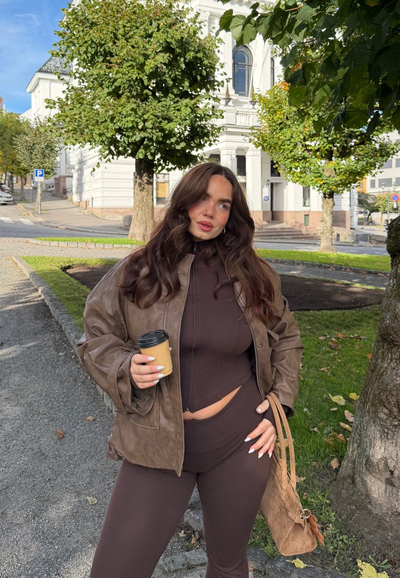 Brun skinnjakke over en matchende tettsittende topp og leggings. Kopp i hånden, omgitt av grøntområder med urbane bygninger i bakgrunnen.