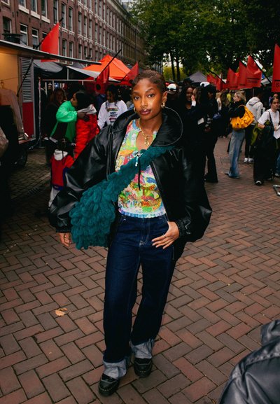 Abrigo puffer negro de gran tamaño, camisa de patrón colorido, jeans azules, zapatos negros gruesos y bolso verde texturizado. Ambiente de mercado al aire libre.