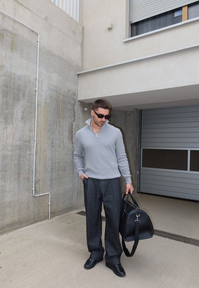 Hombre con suéter gris y pantalones de rayas sostiene un bolso negro de cuero cerca de una pared de hormigón y una puerta de garaje cerrada.