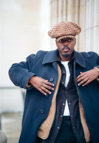 Ναυτικά μπλε παλτό από μαλλί με μεγάλο γιακά, στρωμένο πάνω από ένα μαύρο πουκάμισο με κουμπιά και λευκή μπλούζα. Συνδυασμένο με καπέλο καφέ με σχέδια.