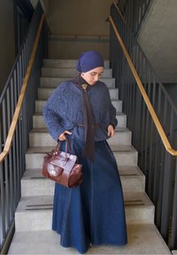 Pull en maille épaisse bleu marine, longue jupe en jean, sac à main en cuir marron. Écharpe nouée autour du col, tissu texturé et détails de conception superposés.