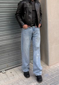 Personne portant une veste en cuir noire, une chemise noire, un jean bleu clair et des chaussures noires, debout sur un trottoir carrelé près d'une grille métallique et d'un mur en pierre.