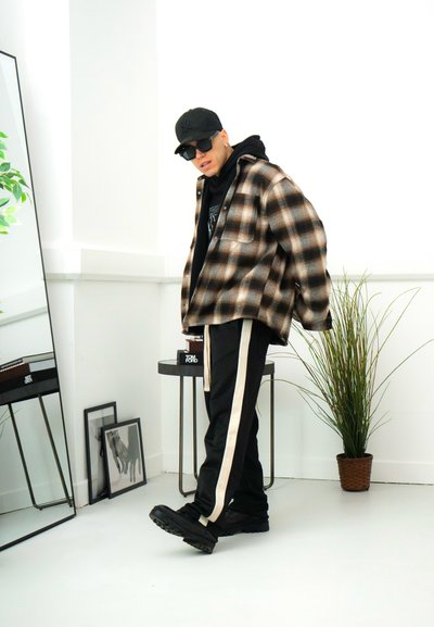 Camisa de franela de cuadros marrones de gran tamaño sobre una sudadera negra con capucha, combinada con pantalones negros con rayas blancas laterales y zapatillas negras gruesas.