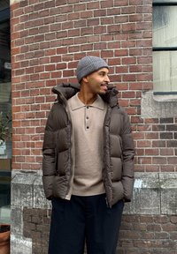 Doudoune marron avec col montant, fermeture éclair et poignets élastiques portée sur un pull beige, associée à un pantalon foncé, devant un mur en briques.