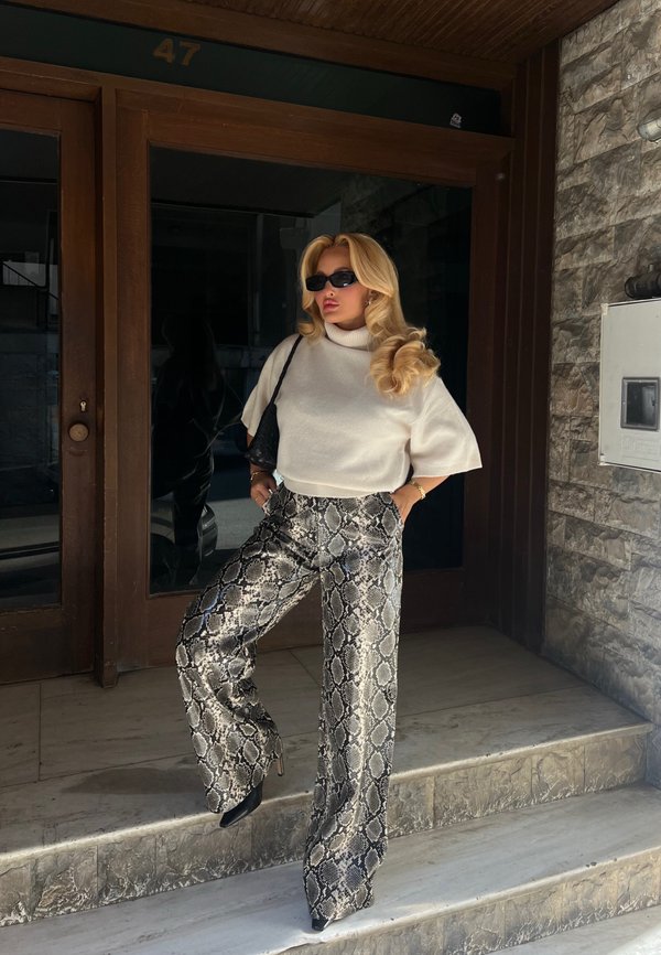 Mujer con cabello rubio que lleva gafas de sol, jersey de cuello alto de manga corta blanco, pantalones con estampado de serpiente, tacones negros y un bolso negro de hombro, parada en unos escalones.