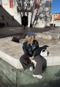 Casaco oversized de couro preto, calças largas cinzentas, mala branca, boné de ganga, sentado na borda de pedra de uma piscina transparente, dia ensolarado.
