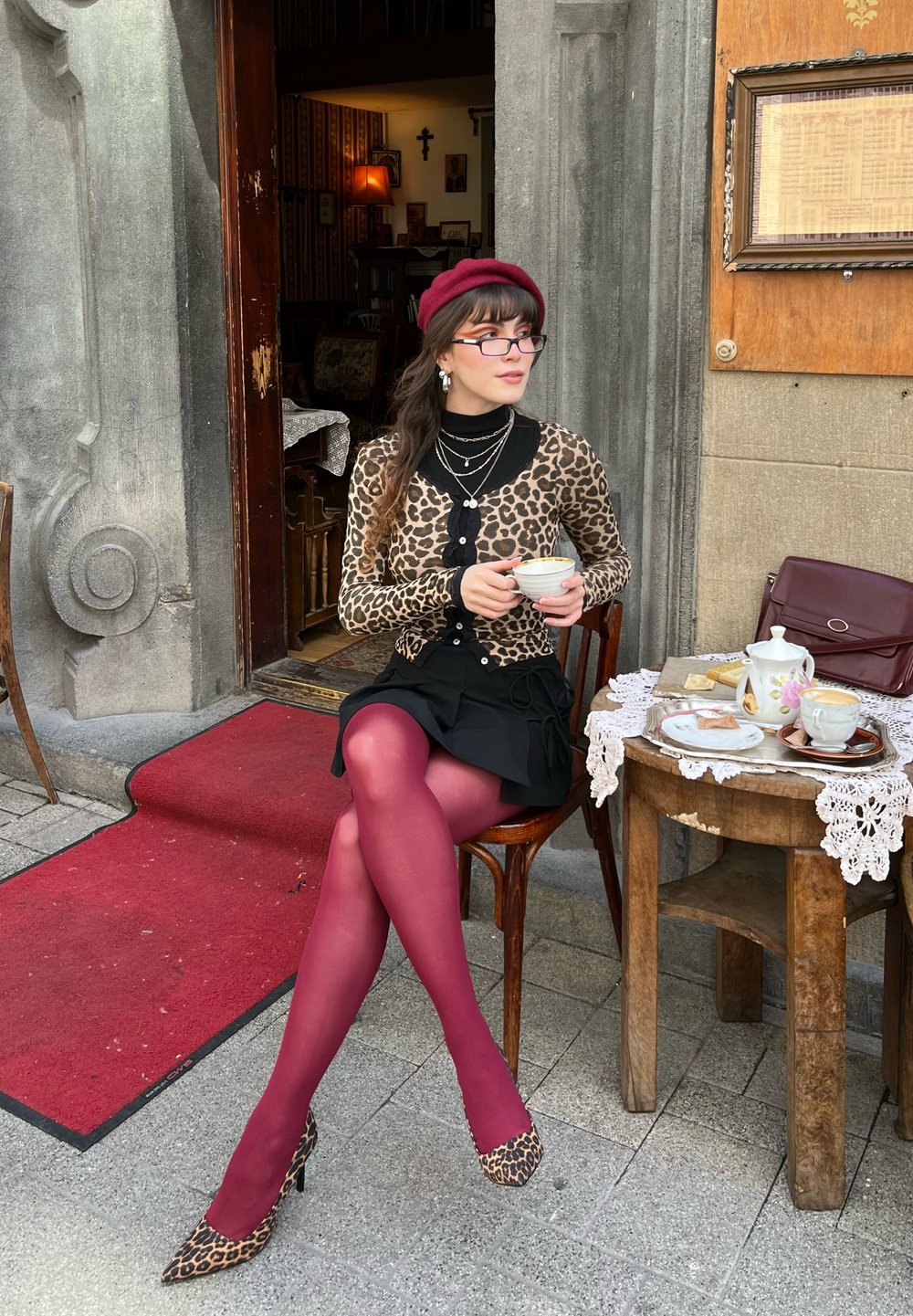 Καρντιγκάν με λεοπάρ σχέδιο, μαύρη φούστα και μπορντό καλσόν. Καθισμένη σε ξύλινο τραπέζι στρωμένο με τσαγιέρες, με ένα διακοσμητικό υφαντό τραπεζομάντιλο.