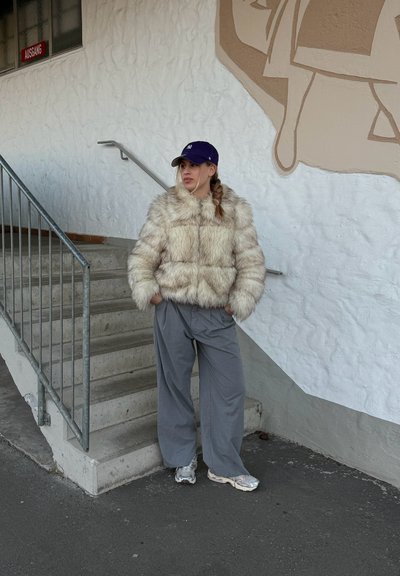 Chaqueta acolchada de piel sintética en color crema con cuello alto, combinada con pantalones sueltos grises y zapatillas plateadas. El fondo presenta una pared con textura.