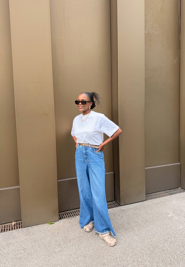 Junge Frau mit Sonnenbrille, weißem T-Shirt, weiten blauen Jeans und beigen Clogs, stehend mit den Händen in der Hüfte vor einer braunen Wand.