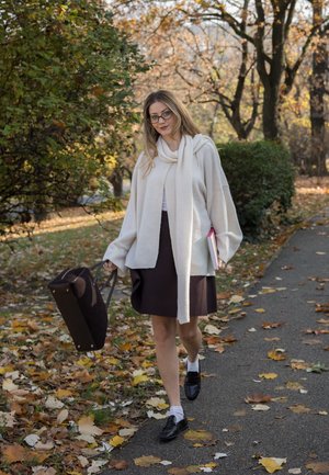 Suéter de crema claro de gran tamaño, falda marrón oscuro, zapatos negros, llevando un bolso oscuro. Hojas de otoño en el suelo, árboles al fondo.
