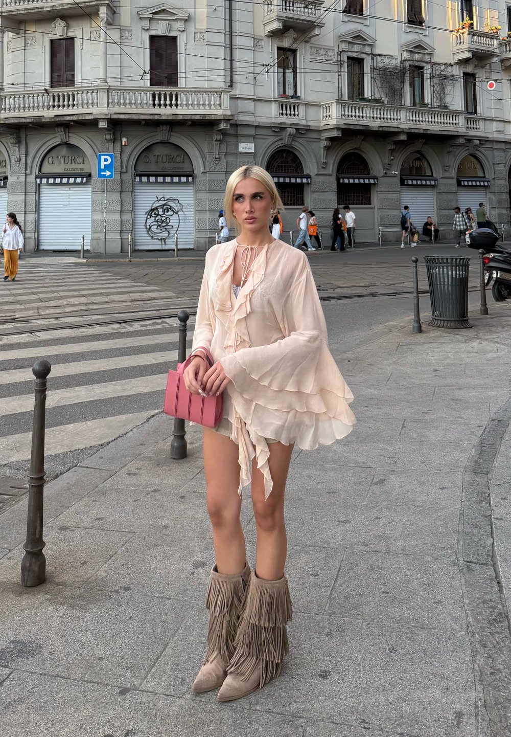 Blusa rizada de color rosa claro con mangas de campana, combinada con botas de flecos beiges y sosteniendo un pequeño bolso rosa. Fondo urbano.