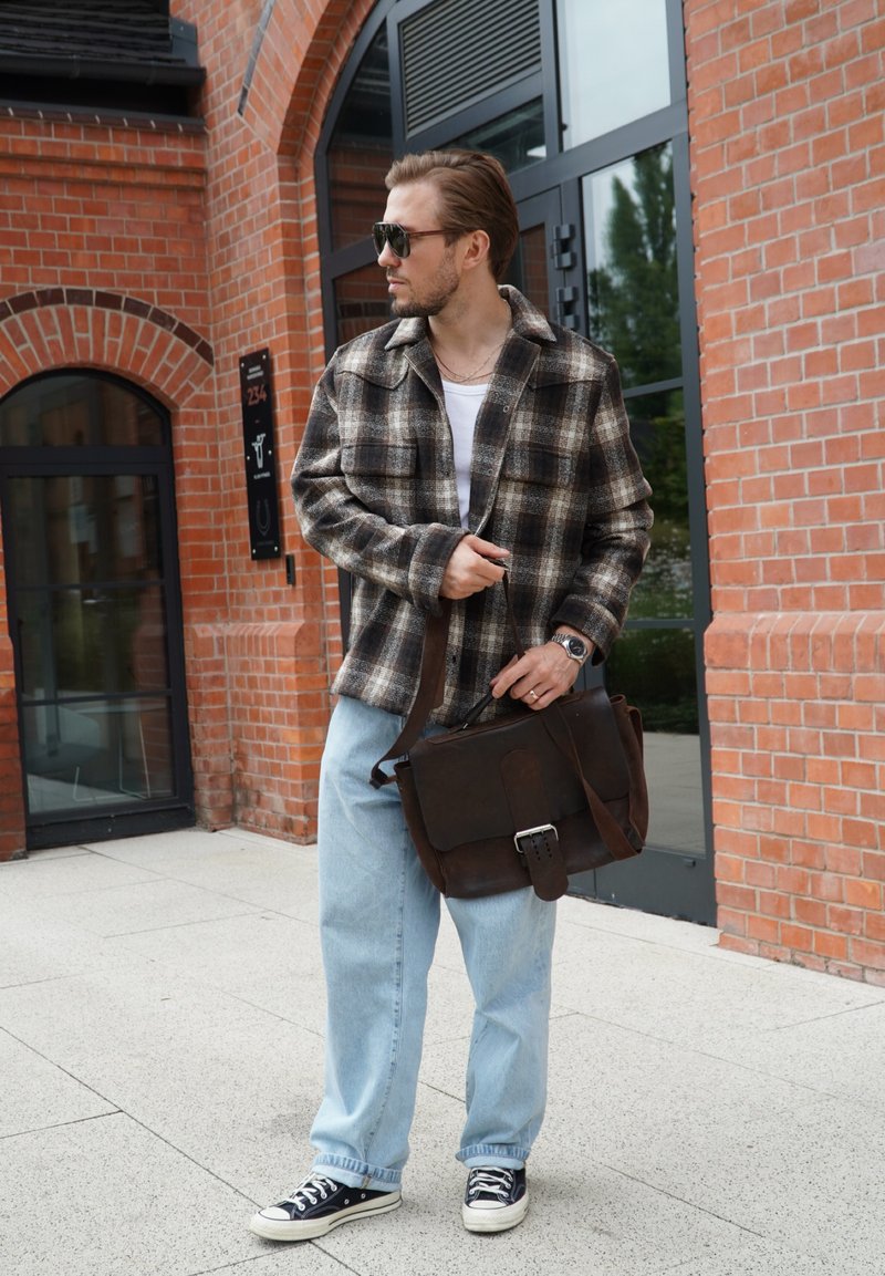 Giacca a scacchi, t-shirt bianca, jeans blu chiaro, sneaker nere. Borsa a spalla in pelle marrone con fibbia. Ambientazione urbana con sfondo di muro in mattoni.