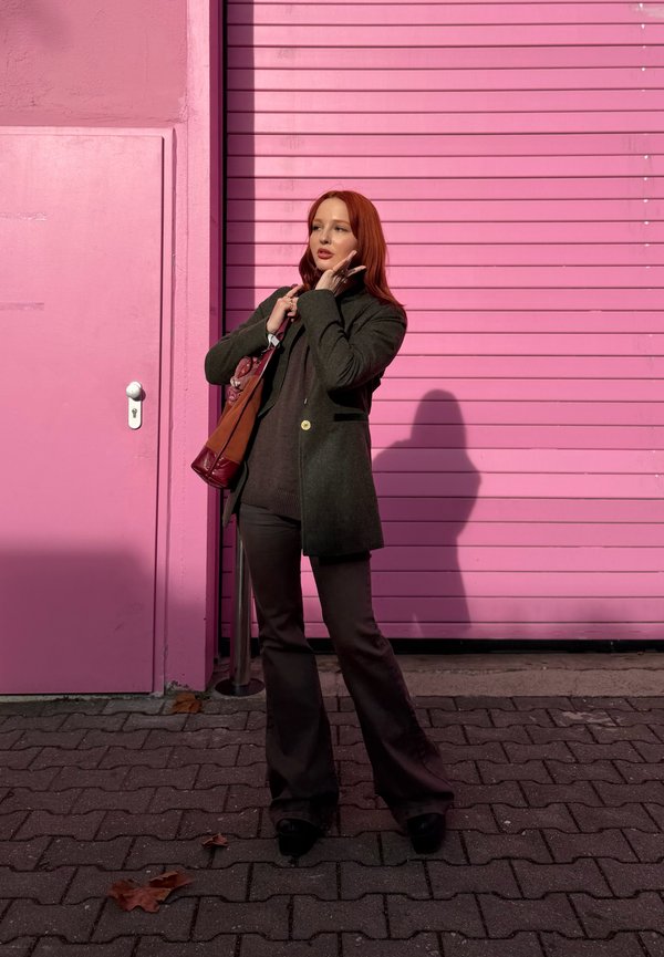 Mlada ženska z rdečimi lasmi, oblečena v temen plašč in hlače, stoji na tlakovanem pločniku ter se dotika obraza, ob svetlo roza vratih in okenski zavesi.