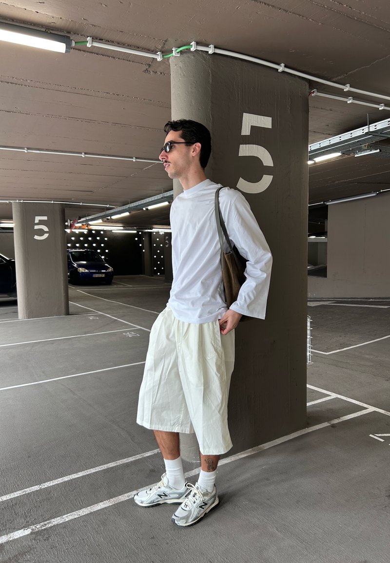 Άνδρας με λευκό πουκάμισο μακρυμάνικο και σορτς, γυαλιά ηλίου και αθλητικά παπούτσια, στέκεται σε κλειστό χώρο στάθμευσης δίπλα σε κολώνα με το νούμερο 5.